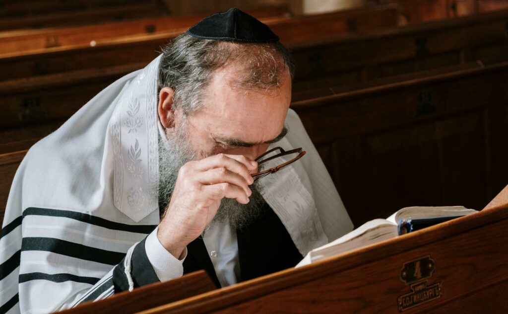 Senior man with a beard and kippah reading a book in a synagogue, adjusting his eyeglasses.