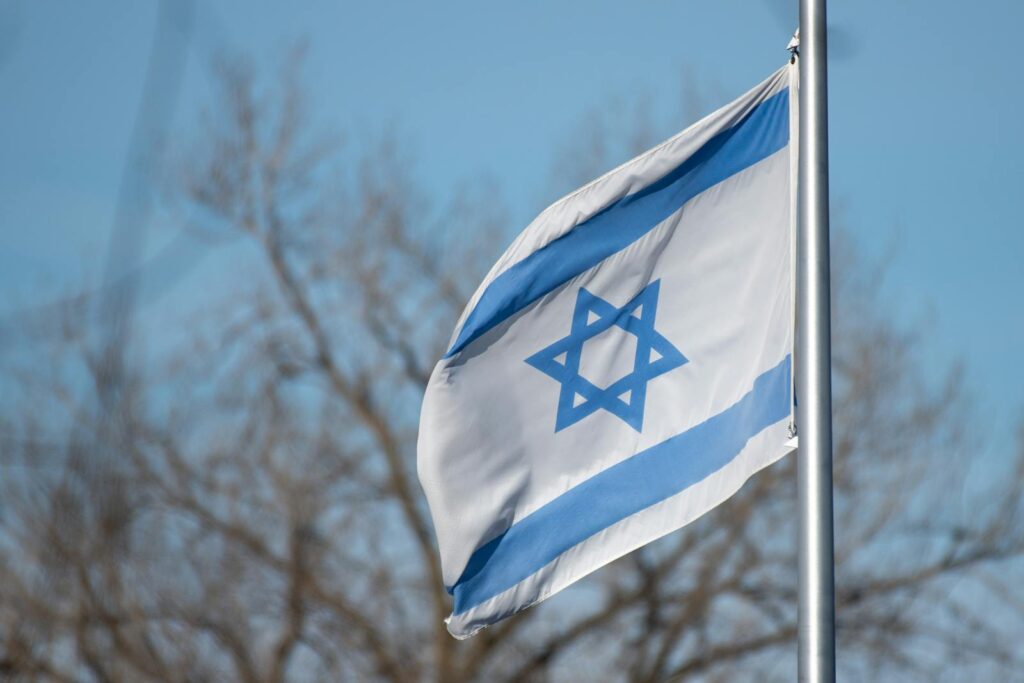 The Israeli flag featuring the Star of David against a clear blue sky and trees.