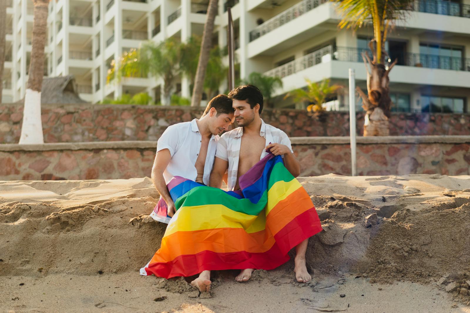 Two men sitting closely on a beach, wrapped in a vibrant rainbow flag, showcasing warmth and intimacy.