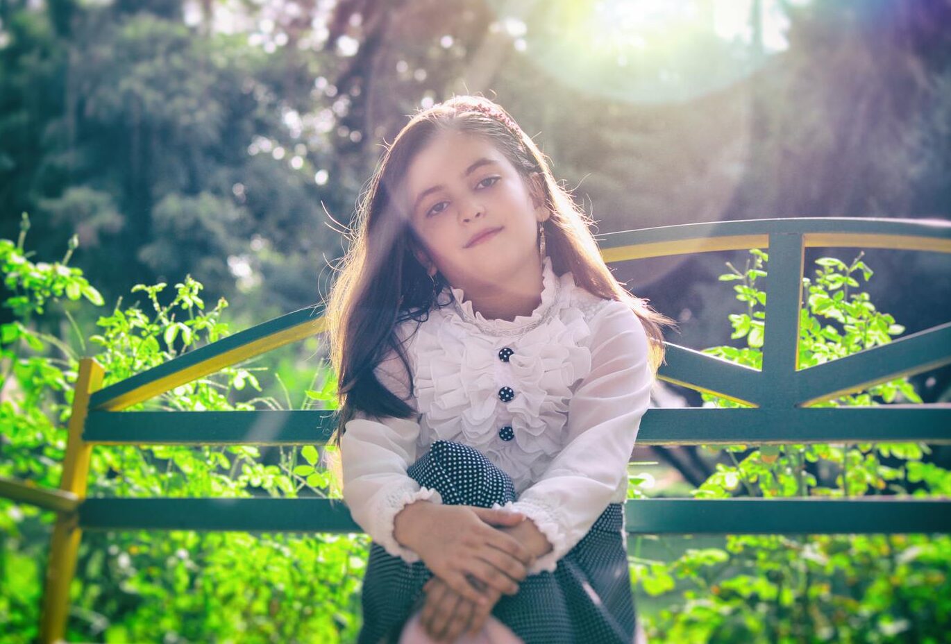 A young girl in stylish attire sitting on a bench in a sunny outdoor garden setting.