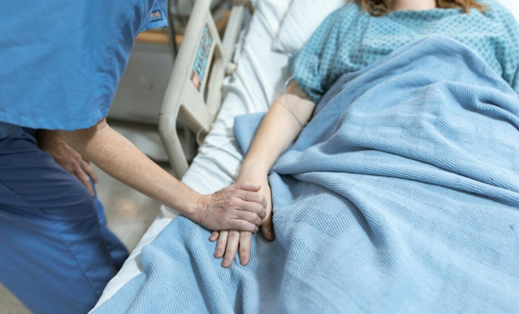 A nurse in scrubs offers comfort by holding a patient's hand in a hospital bed.